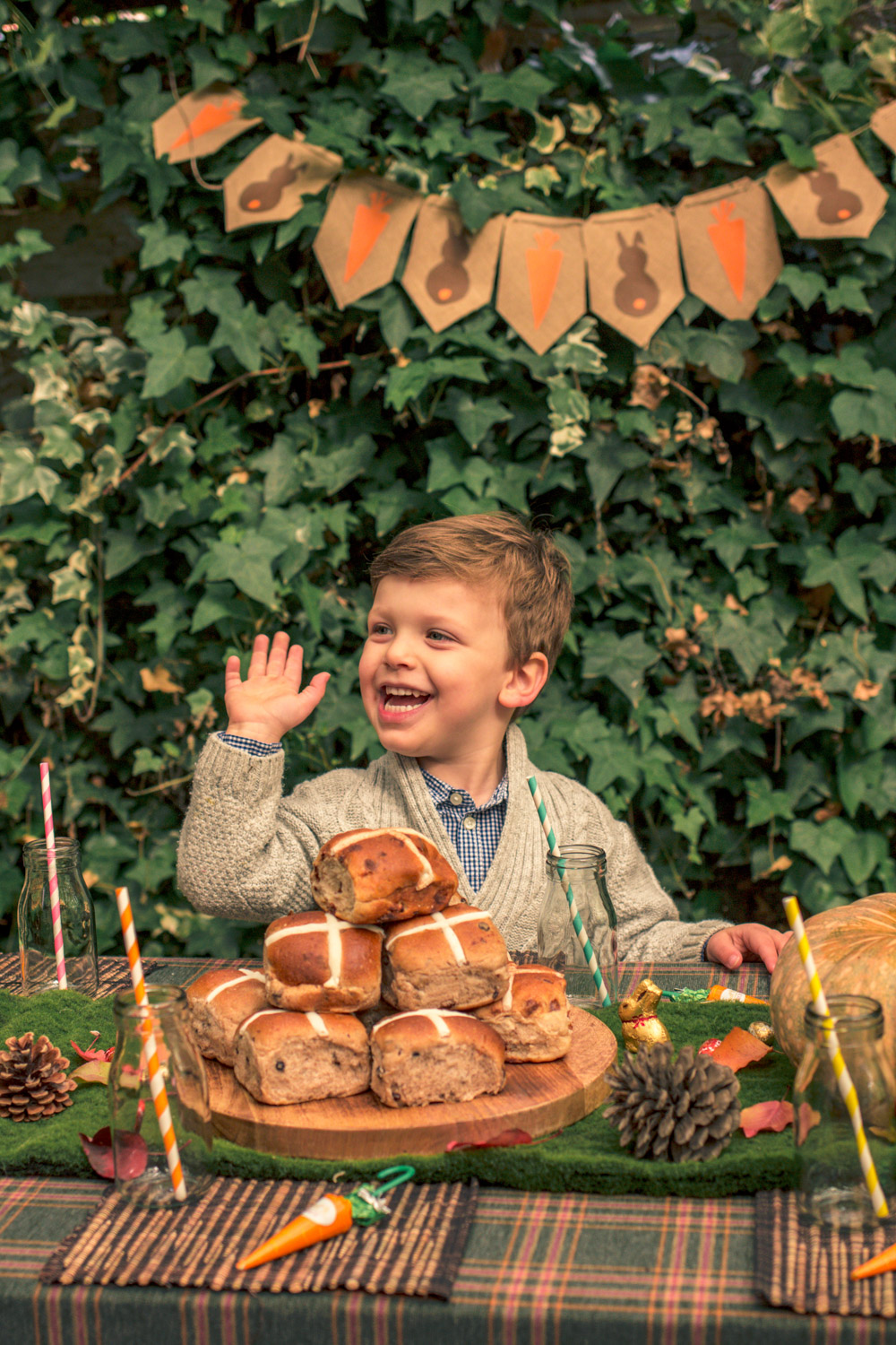 Little boy in Janie and Jack knit cardigan sitting at the kids party table at the Autumn easter egg hunt with hot cross buns and pumpkins and pine cones sitting below autumn themes easter bunting