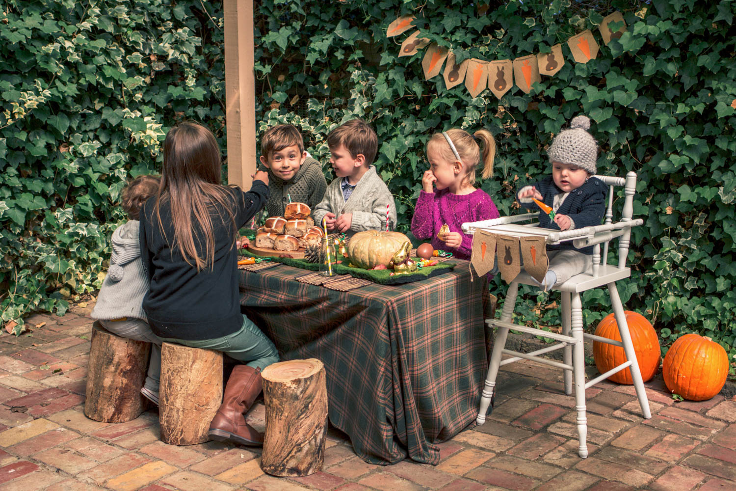 Children seated on wooden logs easting hot cross buns and chocolates at party table for autumn easter egg hunt. Baby seated in vintage wooden high chair with bunting