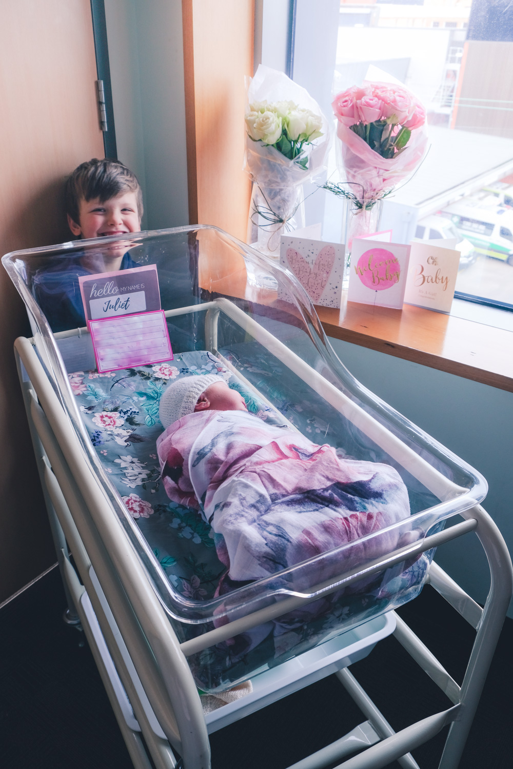 Newborn baby in hospital bed surrounded by flowers and cards and little brother