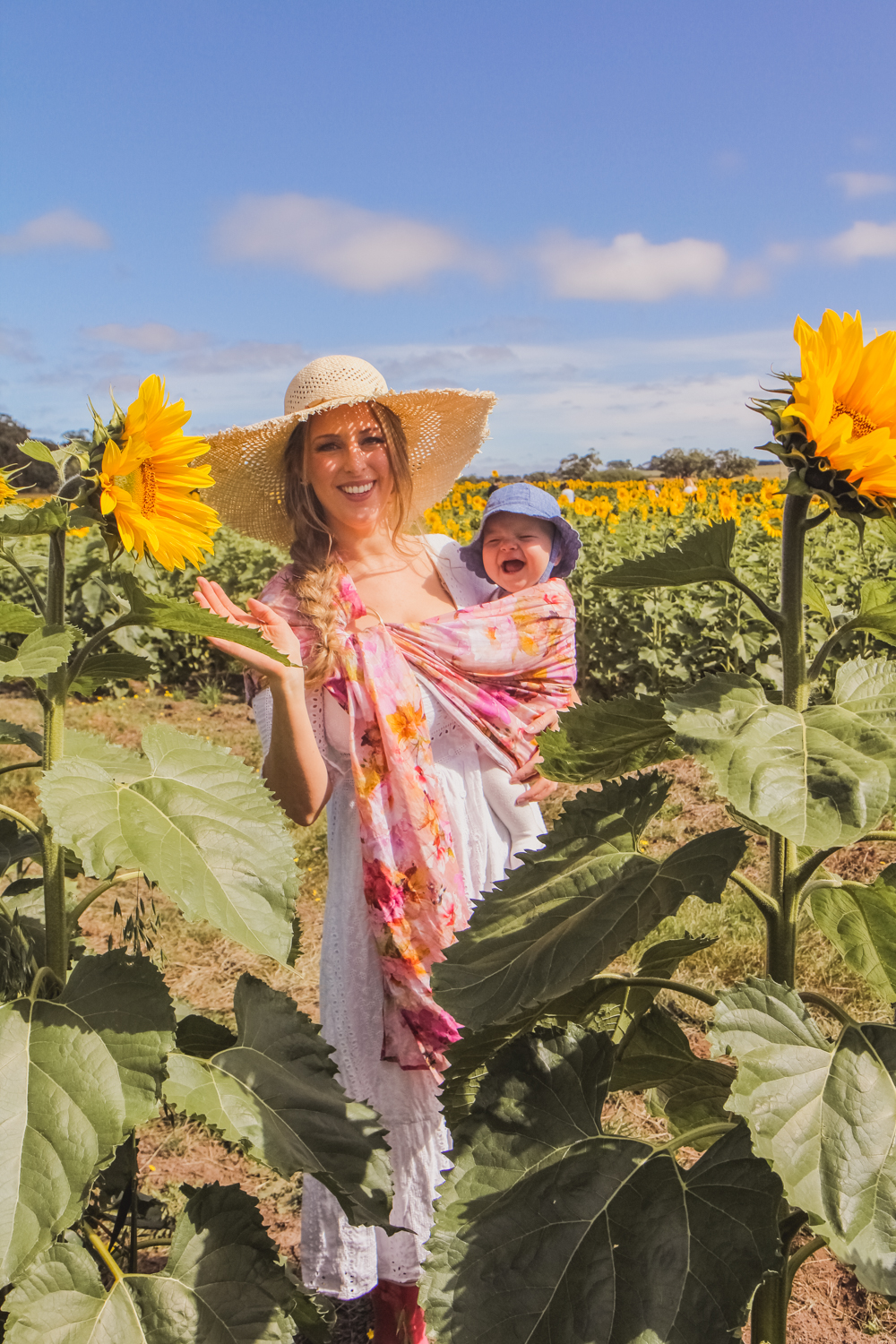 Goldfields Girl at sunflower farm in Ballarat in Victoria. Wearing white eyelet dress, straw hat and ping ring sling baby carrier.