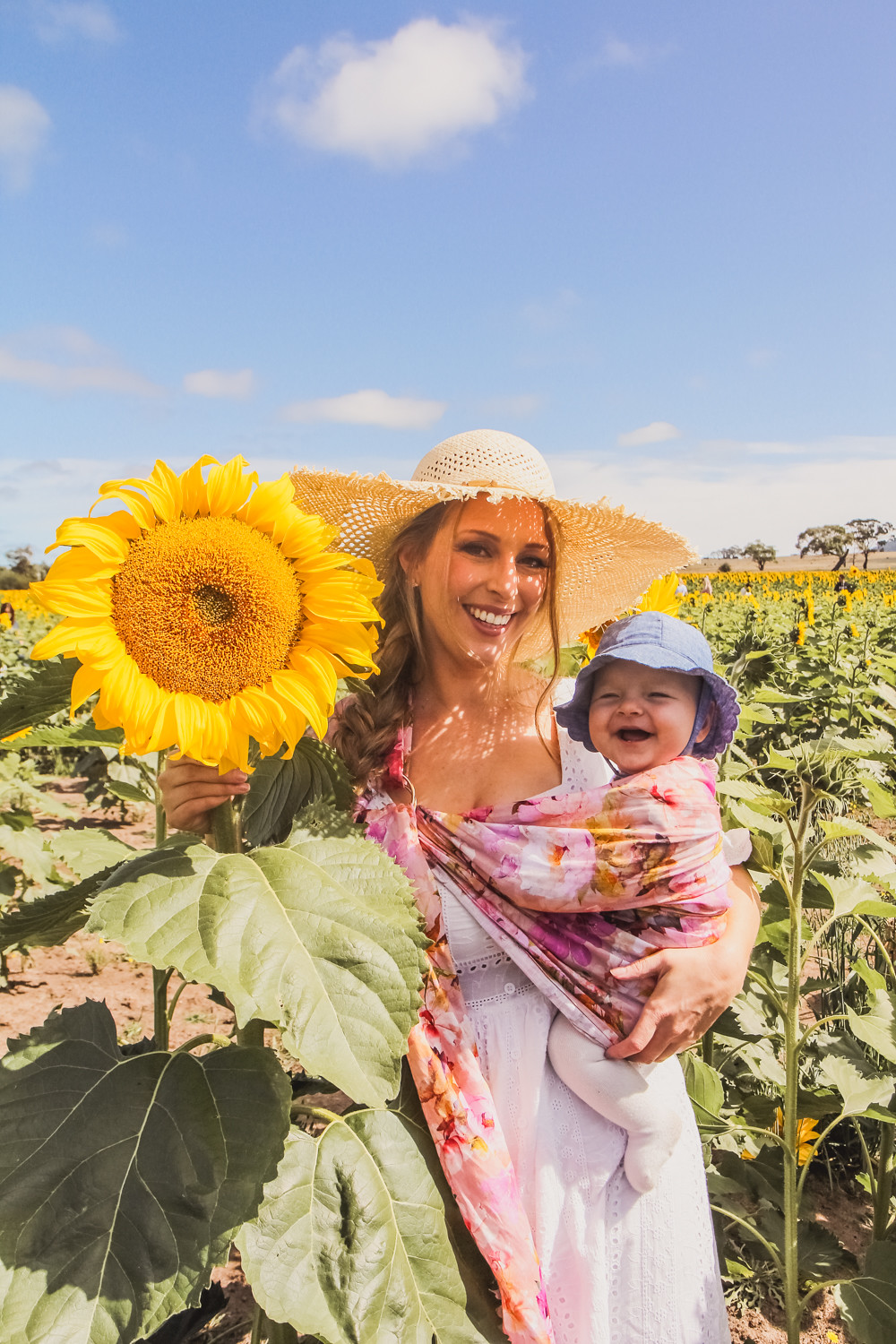 Goldfields Girl at sunflower farm in Ballarat in Victoria. Wearing white eyelet dress, straw hat and ping ring sling baby carrier.