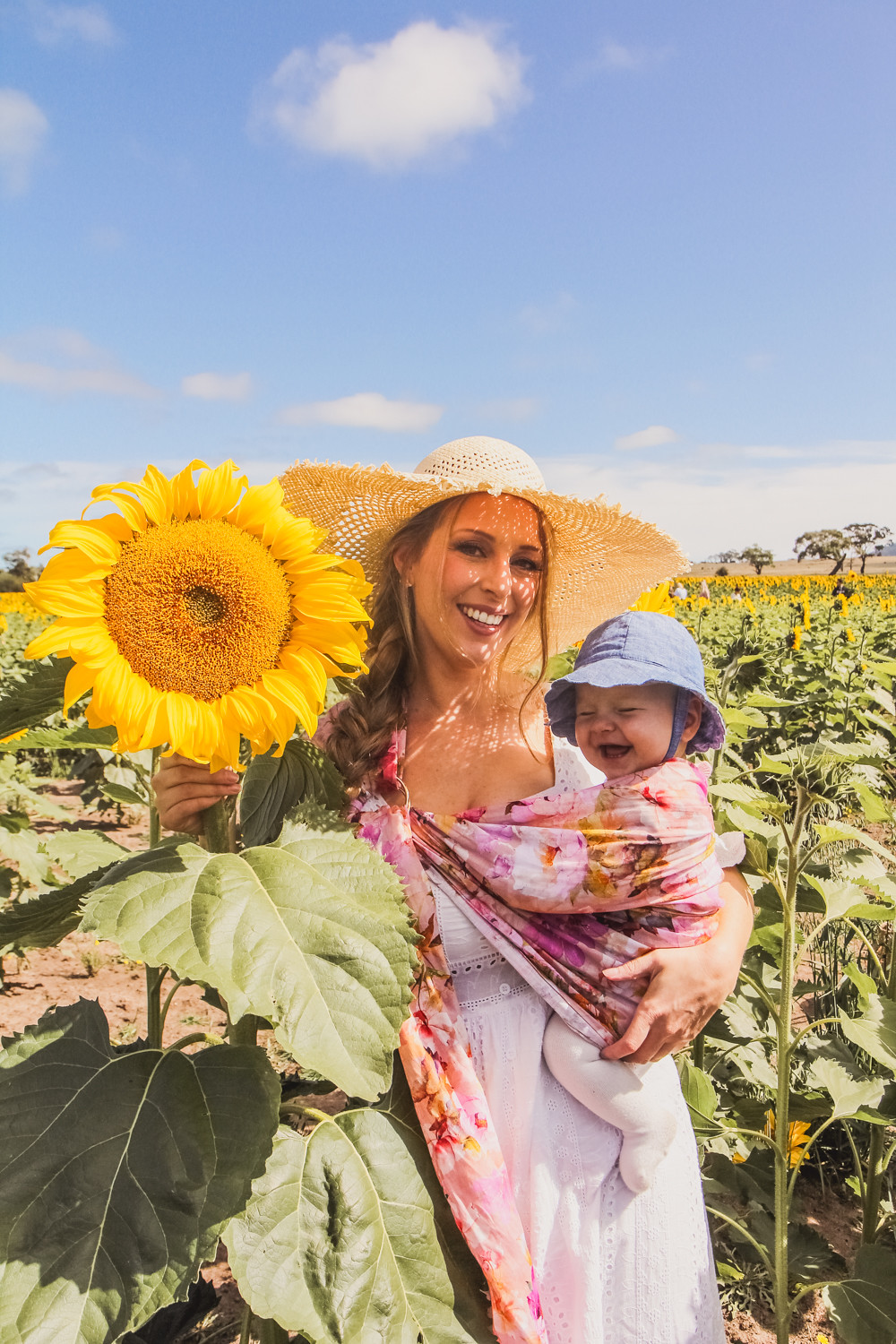 Goldfields Girl at sunflower farm in Ballarat in Victoria. Wearing white eyelet dress, straw hat and ping ring sling baby carrier.