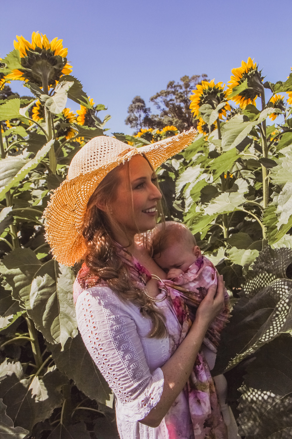 Goldfields Girl at sunflower farm in Ballarat in Victoria. Wearing white eyelet dress, straw hat and ping ring sling baby carrier.