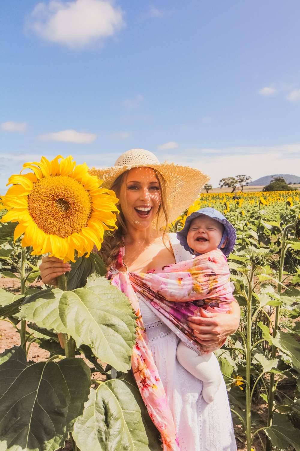 Goldfields Girl at sunflower farm in Ballarat in Victoria. Wearing white eyelet dress, straw hat and ping ring sling baby carrier.