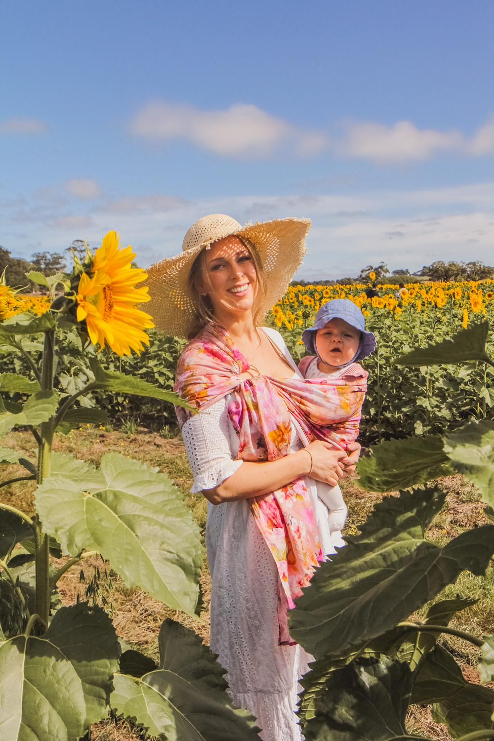 Goldfields Girl at sunflower farm in Ballarat in Victoria. Wearing white eyelet dress, straw hat and ping ring sling baby carrier.