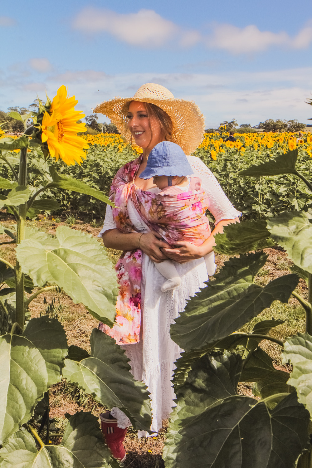 Goldfields Girl at sunflower farm in Ballarat in Victoria. Wearing white eyelet dress, straw hat and ping ring sling baby carrier.