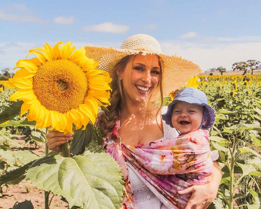 Goldfields Girl at sunflower farm in Ballarat in Victoria. Wearing white eyelet dress, straw hat and ping ring sling baby carrier.