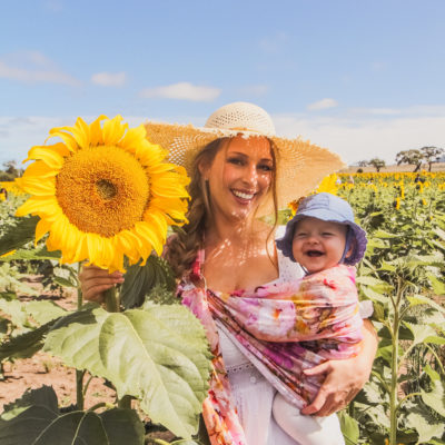 Goldfields Girl at sunflower farm in Ballarat in Victoria. Wearing white eyelet dress, straw hat and ping ring sling baby carrier.